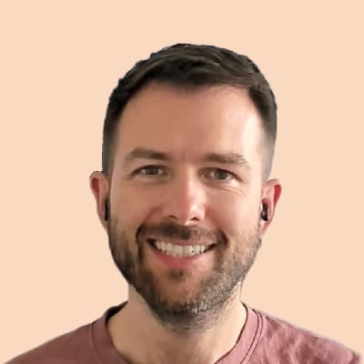 A head-and-shoulders portrait of William, a smiling man with short brown hair, light stubble, and wireless earbuds, wearing a rose-colored crewneck shirt, set against a light peach background.